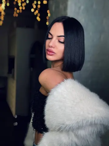 Elegant woman with sleek black bob hairstyle wearing a white faux fur coat in a moody, dimly lit setting.