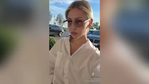 Stylish blonde woman in a white button-down shirt and sunglasses posing outdoors in bright daylight.