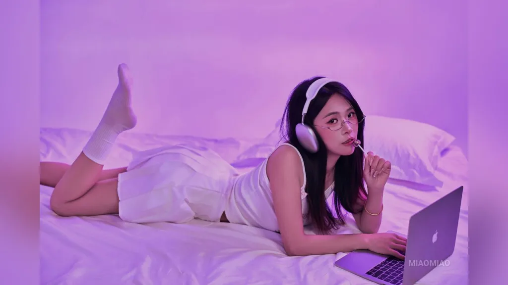 miaomiao — miaomiao lies on a white bed wearing white tank top and shorts while using headphones and a laptop under purple lighting.