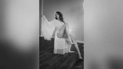 Elegant woman in a white lace flowing gown posing gracefully in a moody black and white indoor setting.