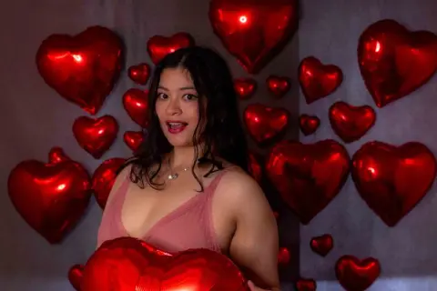 Young woman in a pink lace top smiling amidst red heart balloons in a romantic studio setting.