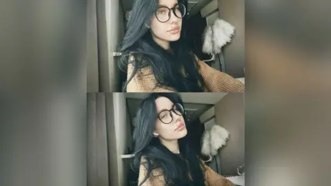 Young woman with long dark hair and glasses wearing a cozy brown sweater in a bright indoor setting.