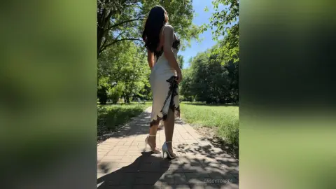 Elegant woman in a white lace slip dress and high heels walking on a sunny park path.