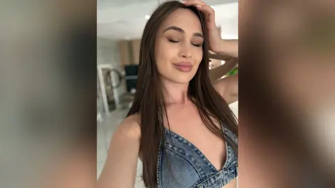 Young woman with long brown hair wearing a denim top in a bright, minimalist indoor setting.