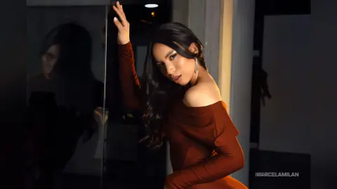 MarcelaMilan poses in a tight brown off-the-shoulder dress with her long dark hair flowing over her shoulder.