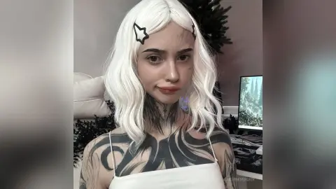 Young woman with white wavy hair and neck tattoos wearing a white camisole in a cozy indoor setting.