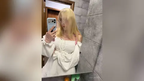 JudiJadore poses for a mirror selfie wearing a white off-the-shoulder top with blonde hair and glasses.