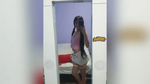 SofiaCorazon poses in a mirror wearing a pink tank top and white skirt, showing off her long dark hair in a bedroom setting.