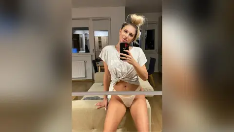 Young woman in a white t-shirt and beige underwear taking a mirror selfie in a modern living room.