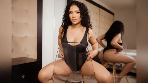 Curvy woman in a black bodysuit sitting on a clear chair in a modern bedroom setting.
