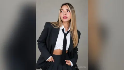 Confident young woman in a black blazer and tie outfit posing against a neutral studio background.