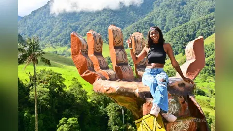 NatyGlow smiles while sitting on a large brown hand sculpture, wearing a black crop top and light blue jeans against a lush green mountain backdrop.