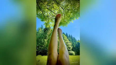 EmaLowee lifts her bare legs and feet high toward the green leaves of a tree against a clear blue sky.
