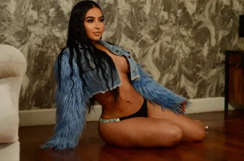 Young woman in a blue faux fur denim jacket sitting on a wooden floor against a textured wall.