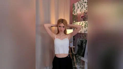 Blonde woman in a white crop top and black pants posing in a cozy room with indoor plants.