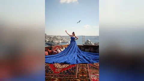 Elegant woman in a flowing royal blue gown posing on a rooftop terrace overlooking a coastal city skyline.