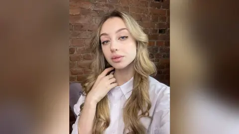 Blonde woman in a white button-down shirt posing with a serene expression against a rustic brick wall.