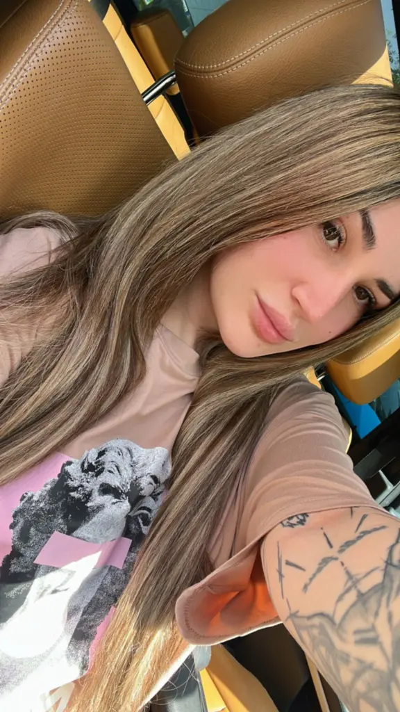 Young woman with long dark hair and tattoos posing in a cozy bedroom setting with a relaxed mood.