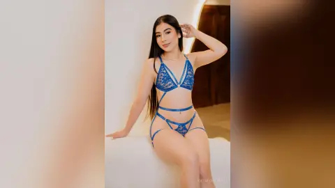 Young woman in blue lace lingerie set sitting on a white surface in a bright indoor setting.