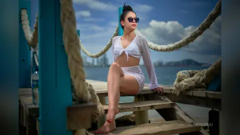 JessieLiu sits on a wooden pier wearing a white tie-front crop top and matching white bottoms, looking off to the side in sunglasses.