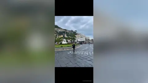 A traveler in dark clothing walking through a scenic coastal town street in Sorrento, Italy under a cloudy sky.