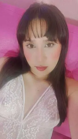 Young woman with dark bangs in a white lace top posing against a pink background.