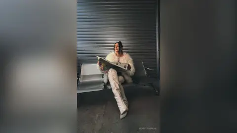 Stylish woman in white faux fur coat and boots reading a newspaper in a moody industrial setting.