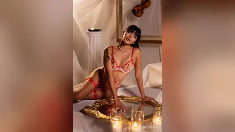 Elegant woman in red lace lingerie posing on a bed in a romantic, dimly lit room with candlelight.