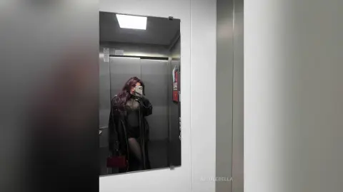 NicoleBella takes a mirror selfie in an elevator wearing a black bodysuit and a long black coat.