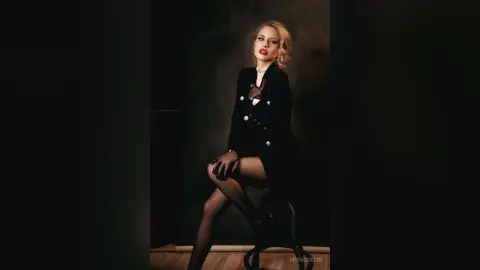 Elegant blonde woman in a black blazer and tights posing in a moody, dark studio setting.