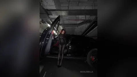 NicoleBella stands in a black car wearing a sheer black bodysuit and black tights.