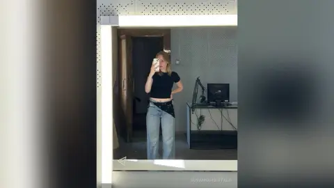 Young woman in black t-shirt and light blue jeans taking a mirror selfie in a modern bedroom.