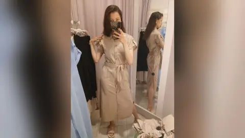 Woman in a beige belted midi dress taking a mirror selfie in a clothing boutique fitting room.
