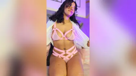 NaomiVelour poses in a pink cut-out bra and matching strap bikini, showing off her dark curly hair and cleavage in a soft-lit room.
