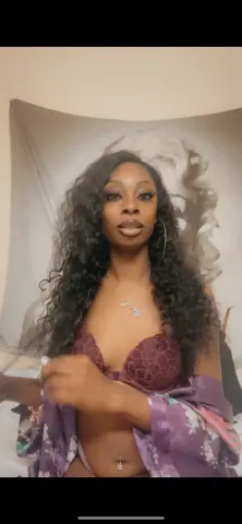 Beautiful woman with long curly hair wearing a purple floral robe and lace lingerie in a cozy bedroom setting.