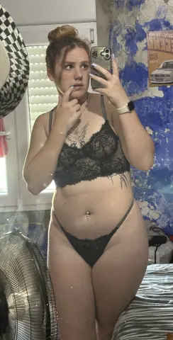 Young woman in black lace lingerie set taking a mirror selfie in a bright bedroom.
