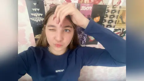 MartineKibbey winks and pouts her lips while wearing a blue long-sleeved sweatshirt, with her hand touching her hair against a wall of music posters.