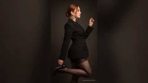 Elegant redhead woman in a black blazer and tights posing confidently against a dark studio background.