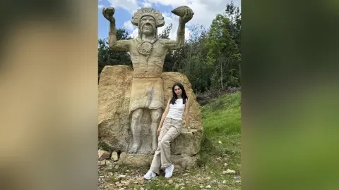 Woman in white tank top and khaki pants standing by a large ancient stone statue in a lush green forest.