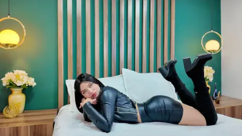 ScarlettPrescott lies on a bed wearing a black leather jacket and a short black leather skirt with black boots, posing with her legs raised against a green wall.