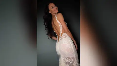 Smiling woman in a white lace backless evening gown posing against a dark studio background.