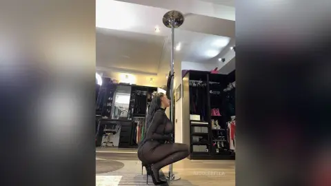 NicoleBella poses in a black bodysuit while kneeling next to a silver dance pole in her studio.