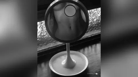 Black and white reflection of a woman in a minimal outfit in a circular vanity mirror on a windowsill.