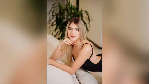 Blonde woman in a black bodysuit relaxing on a white sofa in a bright, minimalist living room.