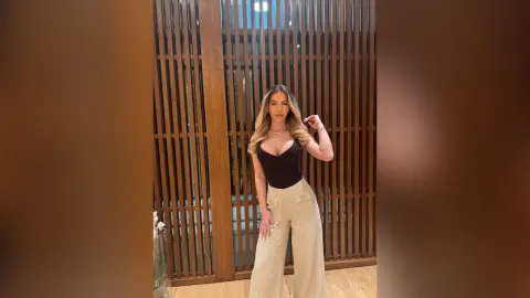 Elegant woman in a black bodysuit and cream wide-leg trousers posing against a modern wooden slat wall.