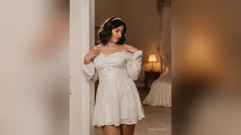 Elegant woman in a white lace cocktail dress posing in a warm, moody bedroom setting.
