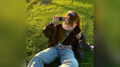 Young woman in black top and jeans taking a photo on a sunny grass field with a vintage camera.