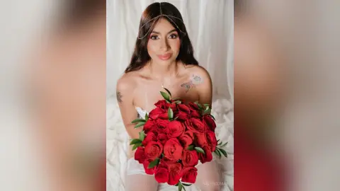 Beautiful woman in white lace holding a large bouquet of red roses in a soft, romantic indoor setting.