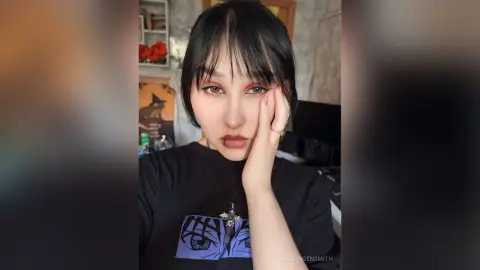 Young woman with dark bangs wearing a black graphic t-shirt in a cozy indoor setting.