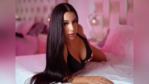 Beautiful woman with long dark hair in a black bodysuit relaxing on a white bed in a soft pink lit bedroom.